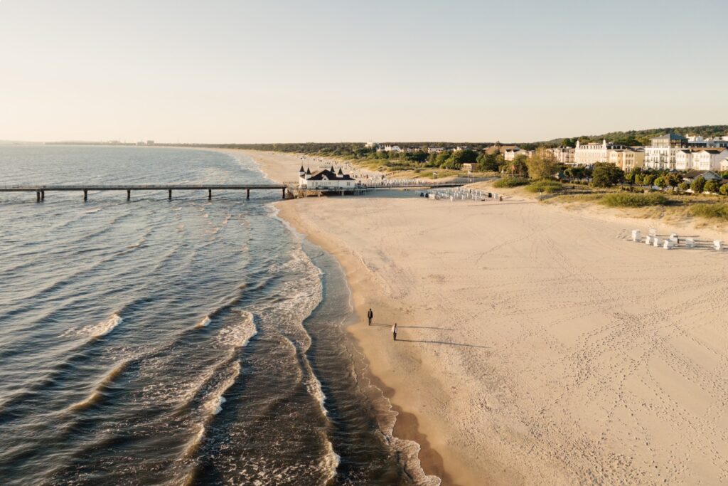 Drone shot of a wide sandy beach stretching to the horizon. To the left, the sea is spanned by a flat bridge from the beach; to the right, a town with bright white facades borders the beach. In the center, two people are walking on the beach.
