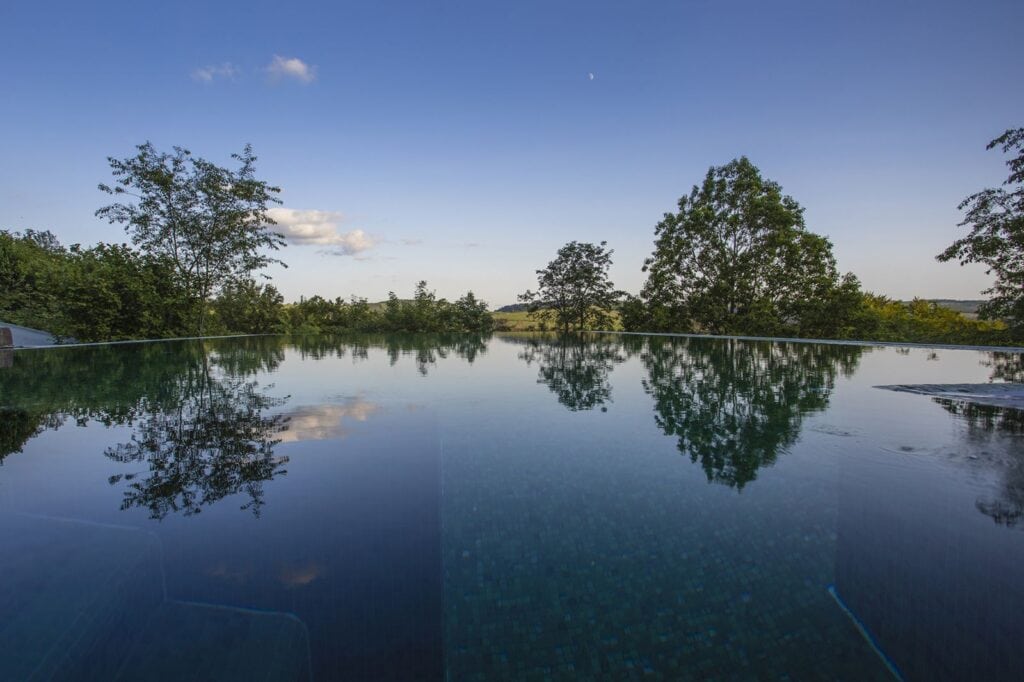 Die untere Hälfte wird durch die Oberfläche eines Infinitypools eingenommen. Hinter dem Beckenrand ragen einige Bäueme hervor, dahinter erkennt man am Horizont einen grün bewaldeten Hang, über allem blauer Himmel