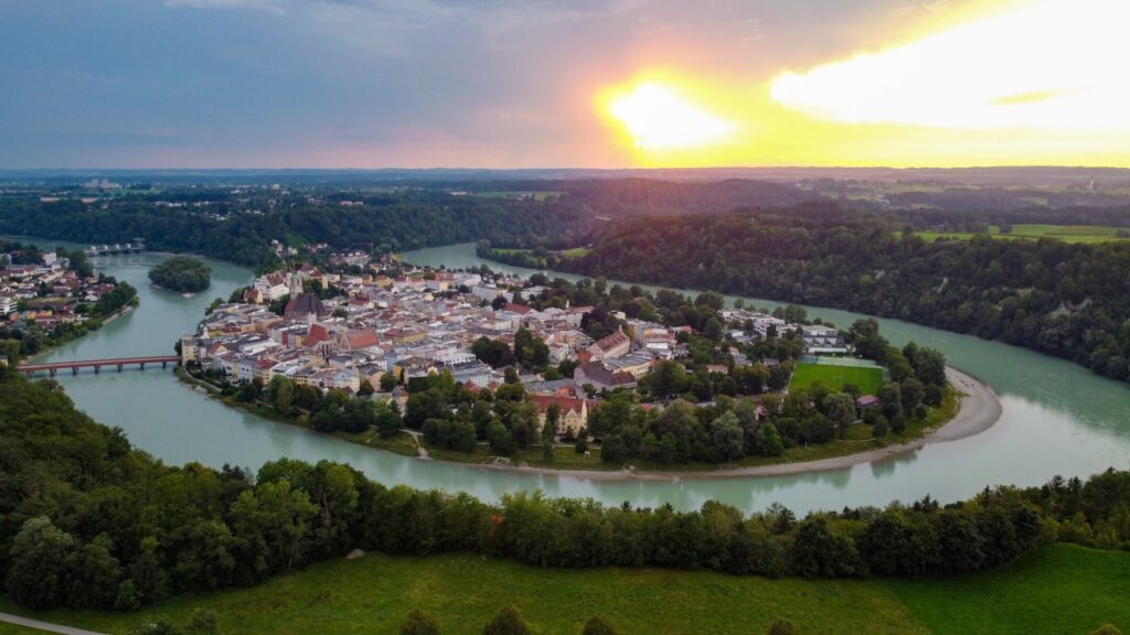 Eine tropfenförmige Insel liegt in einer Flussschleife. Die Insel ist fast vollständig von einer kleinen Stadt ausgefüllt, das Ufer ist jedoch fast rundum von Bäumen bewachsen. Das Gebiet um Fluss und Insel herum ist von Wald bedeckt, dazwischen einzelne Felder. Durch die Wolken bricht das Licht des Sonnenuntergangs.