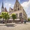 Ein gepflasterter Platz in einer Kleinstadt. Links ragt hinter einem Baum eine weiße Kirche hervor, rechts davon zwei Häuser mit stufigen Giebeln und gotischen Fenstern, die teilweise verkleidet und mit Wappenfiguren bemalt sind. Auf dem Platz sitzen viele Menschen auf den Stühlen einer Außengastronomie. Aus einer Straße auf der rechten Bildseite fahren zwei Radfahrer auf den Betrachter zu.