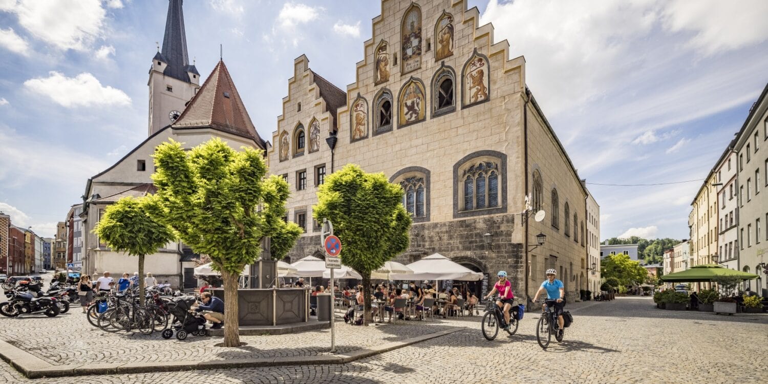 Ein gepflasterter Platz in einer Kleinstadt. Links ragt hinter einem Baum eine weiße Kirche hervor, rechts davon zwei Häuser mit stufigen Giebeln und gotischen Fenstern, die teilweise verkleidet und mit Wappenfiguren bemalt sind. Auf dem Platz sitzen viele Menschen auf den Stühlen einer Außengastronomie. Aus einer Straße auf der rechten Bildseite fahren zwei Radfahrer auf den Betrachter zu.