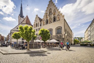 Ein gepflasterter Platz in einer Kleinstadt. Links ragt hinter einem Baum eine weiße Kirche hervor, rechts davon zwei Häuser mit stufigen Giebeln und gotischen Fenstern, die teilweise verkleidet und mit Wappenfiguren bemalt sind. Auf dem Platz sitzen viele Menschen auf den Stühlen einer Außengastronomie. Aus einer Straße auf der rechten Bildseite fahren zwei Radfahrer auf den Betrachter zu.