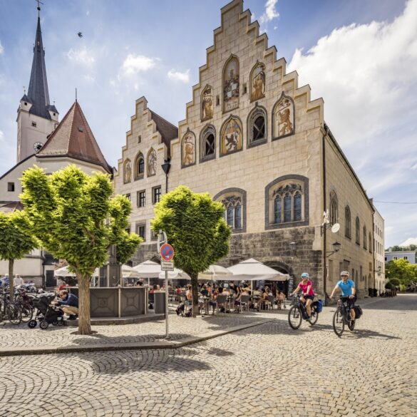 Ein gepflasterter Platz in einer Kleinstadt. Links ragt hinter einem Baum eine weiße Kirche hervor, rechts davon zwei Häuser mit stufigen Giebeln und gotischen Fenstern, die teilweise verkleidet und mit Wappenfiguren bemalt sind. Auf dem Platz sitzen viele Menschen auf den Stühlen einer Außengastronomie. Aus einer Straße auf der rechten Bildseite fahren zwei Radfahrer auf den Betrachter zu.