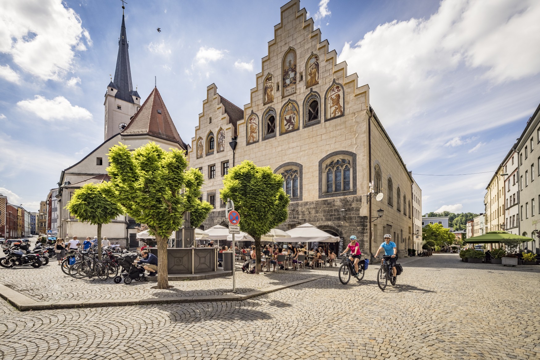 Ein gepflasterter Platz in einer Kleinstadt. Links ragt hinter einem Baum eine weiße Kirche hervor, rechts davon zwei Häuser mit stufigen Giebeln und gotischen Fenstern, die teilweise verkleidet und mit Wappenfiguren bemalt sind. Auf dem Platz sitzen viele Menschen auf den Stühlen einer Außengastronomie. Aus einer Straße auf der rechten Bildseite fahren zwei Radfahrer auf den Betrachter zu.