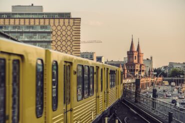 Eine gelbe Straßenbahn fährt über Hochgleis in Richtung einer Brücke mit prominenten, leicht rötlichem doppelten Brückenkopf, markiert durch zwei Türmie mit rundem Aufsatz. Links hinter der Bahn ragen zwei moderne Blockbauten in die Höhe, rechts des Hochgleises eine mehrspurige Straße.