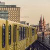 A yellow tram travels along an elevated track toward a bridge with a prominent, slightly reddish double bridgehead, marked by two towers with round tops. To the left behind the tram, two modern block buildings rise up, and to the right of the elevated track is a multi-lane road.