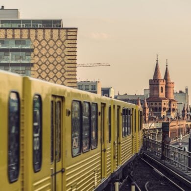 A yellow tram travels along an elevated track toward a bridge with a prominent, slightly reddish double bridgehead, marked by two towers with round tops. To the left behind the tram, two modern block buildings rise up, and to the right of the elevated track is a multi-lane road.