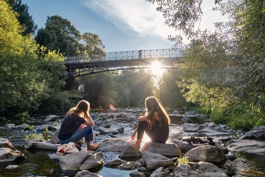 Zwei junge Frauen sitzen auf einigen Steinen in einem flachen Fluss; links und rechts am Ufer stehen einige Bäume, hinter ihnen spannt eine Fußgängerbrücke aus Metall über das Flusstal, durch die die Sonne auf die Szenerie scheint.