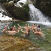 Acht Menschen schwimmen in einem Bergsee und halten sich dabei im Kreis an den Händen. Das Ufer im Hintergrund ist schneebedeckt, rechts endet ein kleiner Wasserfall im See.