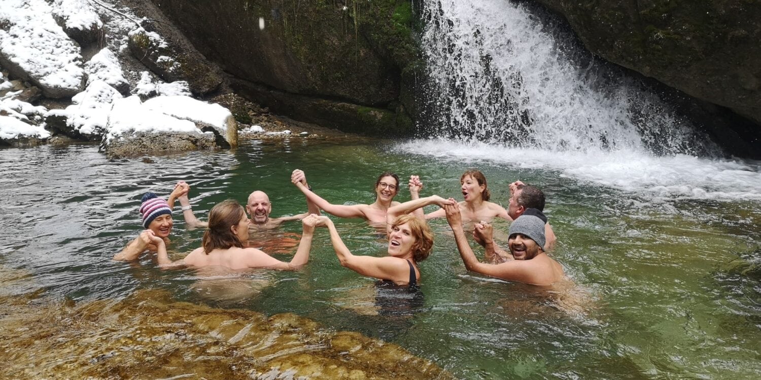 Acht Menschen schwimmen in einem Bergsee und halten sich dabei im Kreis an den Händen. Das Ufer im Hintergrund ist schneebedeckt, rechts endet ein kleiner Wasserfall im See.