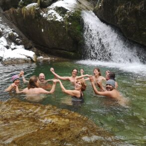 Acht Menschen schwimmen in einem Bergsee und halten sich dabei im Kreis an den Händen. Das Ufer im Hintergrund ist schneebedeckt, rechts endet ein kleiner Wasserfall im See.