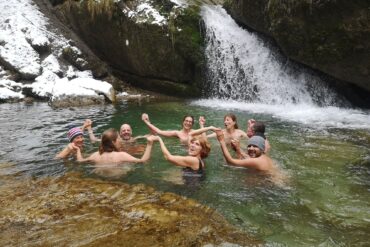 Acht Menschen schwimmen in einem Bergsee und halten sich dabei im Kreis an den Händen. Das Ufer im Hintergrund ist schneebedeckt, rechts endet ein kleiner Wasserfall im See.