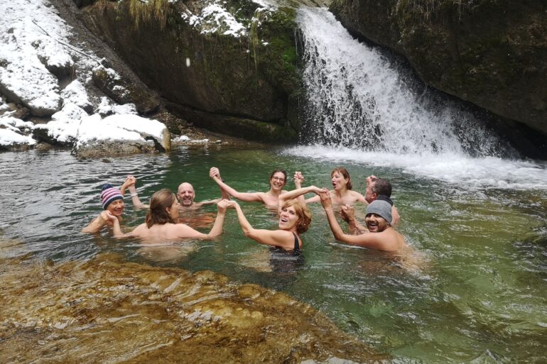 Acht Menschen schwimmen in einem Bergsee und halten sich dabei im Kreis an den Händen. Das Ufer im Hintergrund ist schneebedeckt, rechts endet ein kleiner Wasserfall im See.