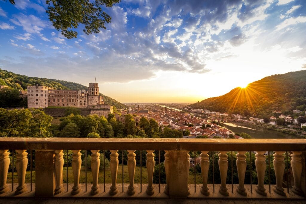 Hinter einem steinernen Barockgeländer erhebt sich zur Linken die Heidelberger Schlossruine, rechts davon im Tal die Altstadt Heidelbergs, rechts daneben der Neckar. Wiederum rechts davon ein bewaldeter Berg, hinter dem die Sonne untergeht und alles in ein gold-gelbes Licht taucht.