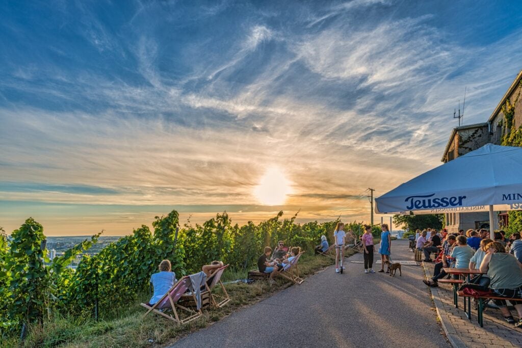 Ein schmaler Asphaltweg führt vom Betrachter zum Horizont; links davon das obere Ende eines Weinbergs, einige Menschen sitzen auf hölzernen Liegestühlen. Rechts vom Weg der Außenbereich einer Bar, über einigen Bierzeltgarnituren ist ein Sonnenschirm gespannt. Die meisten Bänke sind besetzt. Am blauen Himmel geht die Sonne unter.