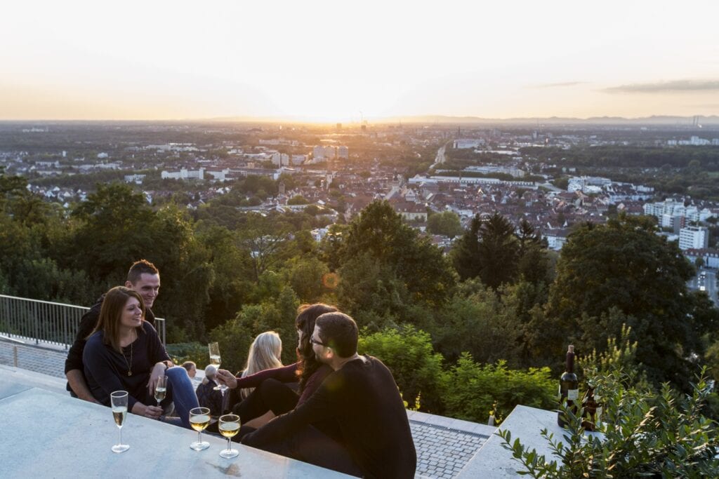 Zwei Pärchen sitzen gemeinsam mit einigen Gläsern Wein auf einer steinernen Terrasse und sind im Gespräch vertieft. Hinter der Terrasse öffnet sich der Blick auf einige Bäume, dahinter ein Tal, das völlig von einer Stadt bedeckt ist. Am Horizont hinter der Stadt geht die Sonne unter.