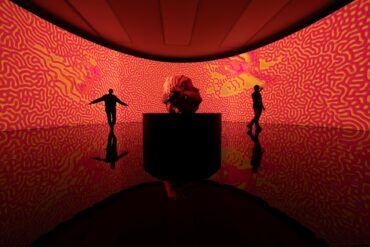 Vor einem riesigen Bildschirm mit rot-gelben Linien steht eine gut zwei Meter hohe Skulptur eines Kopfes, die scheinbar mit einem 3D-Drucker hergestellt wurde. Links und rechts der Skulptur zeichen sich die Silhouetten zweier Personen ab.