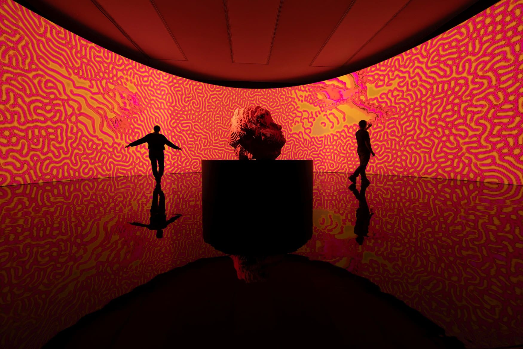 Vor einem riesigen Bildschirm mit rot-gelben Linien steht eine gut zwei Meter hohe Skulptur eines Kopfes, die scheinbar mit einem 3D-Drucker hergestellt wurde. Links und rechts der Skulptur zeichen sich die Silhouetten zweier Personen ab.