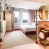 Ein helles Hotelzimmer, von der Tür gesehen; links im Bild ein kleines Wandregal, auf dem unter anderem ein Miniatur-Trabant steht. Rechts ein hinter einem halbhohen Raumtrenner angebrachter Waschtisch mit rundem Becken und schwarzer Armatur, rechts daneben halb zu erkennen der Eingang zum WC. Hinter dem Raumtrenner, auf dem ein großer, runder Spiegel angebracht ist, steht ein breites Bett mit weißen Bezügen. Der Boden ist mit Teppich ausgelegt, die Wände in leichtem Grün gehalten. Hinter dem Bett, auf der dem Betrachter gegenüber liegenden Seite, sind zwei Fenster eingelassen und erhellen den Raum mit Tageslicht.