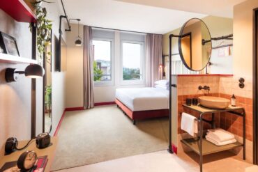 Ein helles Hotelzimmer, von der Tür gesehen; links im Bild ein kleines Wandregal, auf dem unter anderem ein Miniatur-Trabant steht. Rechts ein hinter einem halbhohen Raumtrenner angebrachter Waschtisch mit rundem Becken und schwarzer Armatur, rechts daneben halb zu erkennen der Eingang zum WC. Hinter dem Raumtrenner, auf dem ein großer, runder Spiegel angebracht ist, steht ein breites Bett mit weißen Bezügen. Der Boden ist mit Teppich ausgelegt, die Wände in leichtem Grün gehalten. Hinter dem Bett, auf der dem Betrachter gegenüber liegenden Seite, sind zwei Fenster eingelassen und erhellen den Raum mit Tageslicht.