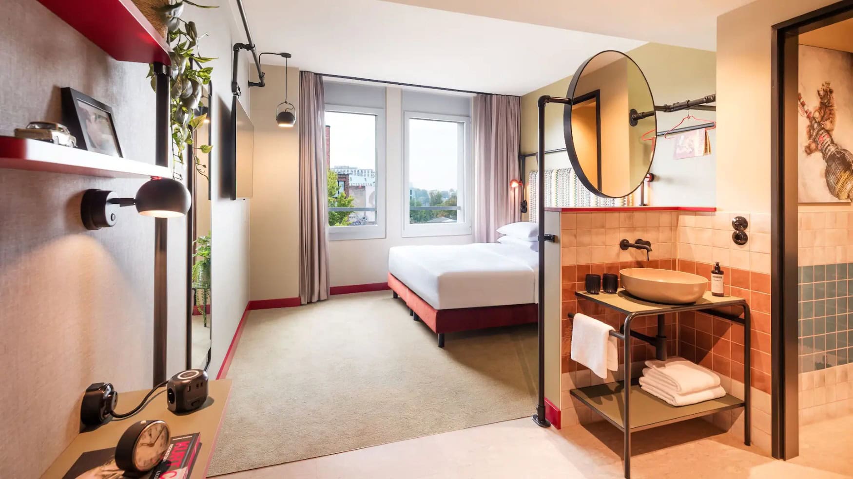 Ein helles Hotelzimmer, von der Tür gesehen; links im Bild ein kleines Wandregal, auf dem unter anderem ein Miniatur-Trabant steht. Rechts ein hinter einem halbhohen Raumtrenner angebrachter Waschtisch mit rundem Becken und schwarzer Armatur, rechts daneben halb zu erkennen der Eingang zum WC. Hinter dem Raumtrenner, auf dem ein großer, runder Spiegel angebracht ist, steht ein breites Bett mit weißen Bezügen. Der Boden ist mit Teppich ausgelegt, die Wände in leichtem Grün gehalten. Hinter dem Bett, auf der dem Betrachter gegenüber liegenden Seite, sind zwei Fenster eingelassen und erhellen den Raum mit Tageslicht.