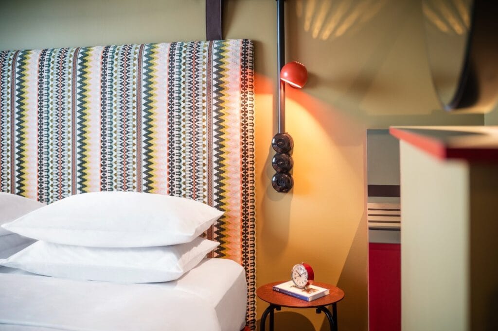 Detailaufnahme eines Hotelbetts; hinter der Matratze mit weißem Bettzeug ist ein Kopfteil mit buntem Webmuster angebracht, rechts davon eine kleine, runde, orangene Wandleuchte, darunter zwei runde, schwarze Lichtschalter und eine ähnlich gestaltete Steckdose. Auf dem Nachttisch steht ein roter Analogwecker. Rechts im Bild ist unscharf ein weißer Raumtrenner erkennbar.