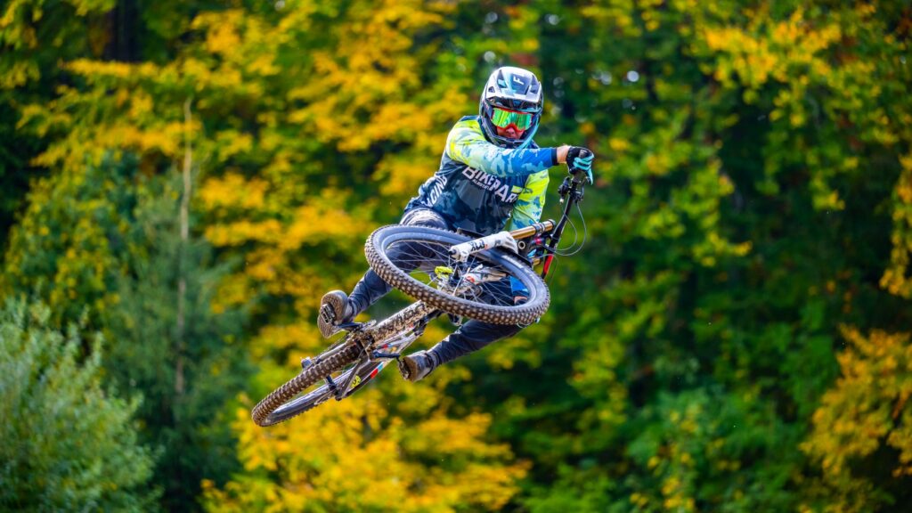 A person in bright cycling gear with a helmet and glasses jumps up on a mountain bike toward the camera, the handlebars pulled to the side. Behind them is a green and yellow forest.