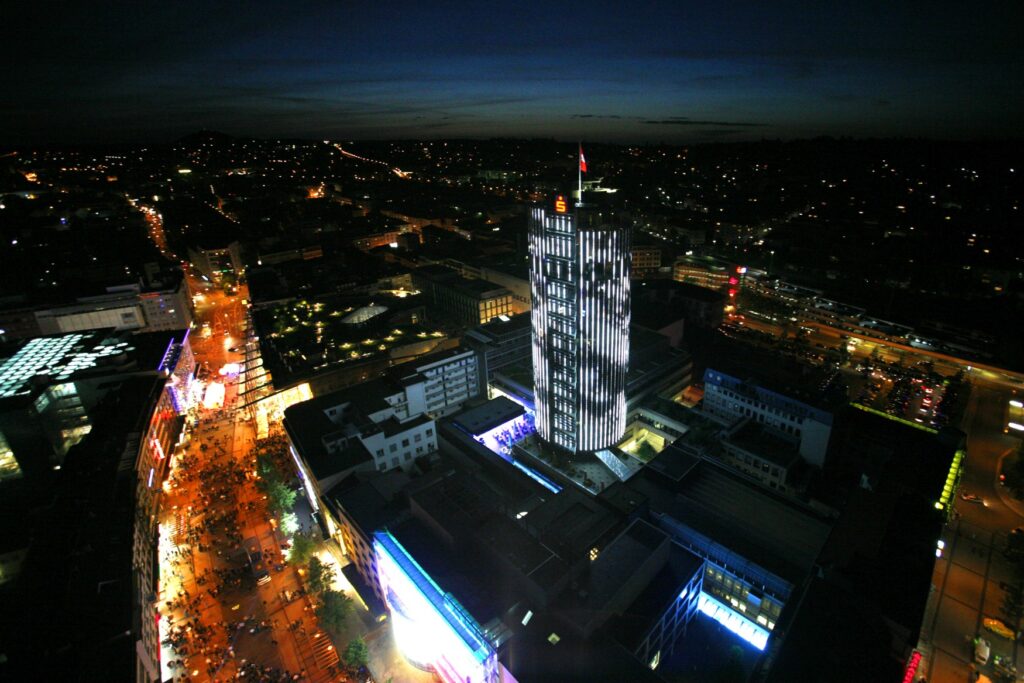 Drohnenaufnahme eines runden Hochauses bei Nacht. Viele Fenster sind erleuchtet, auf dem Dach prangt groß ein rot leuchtendes Sparkassen-Logo. Links vom Gebäude führt eine Fußgängerzone mit vielen Menschen im Schein der Laternen entlang. Im Hintergrund vereinzelt die Lichter einer Stadt. Am Horizont ist noch ganz schwach die Abenddämmerung erkennbar.
