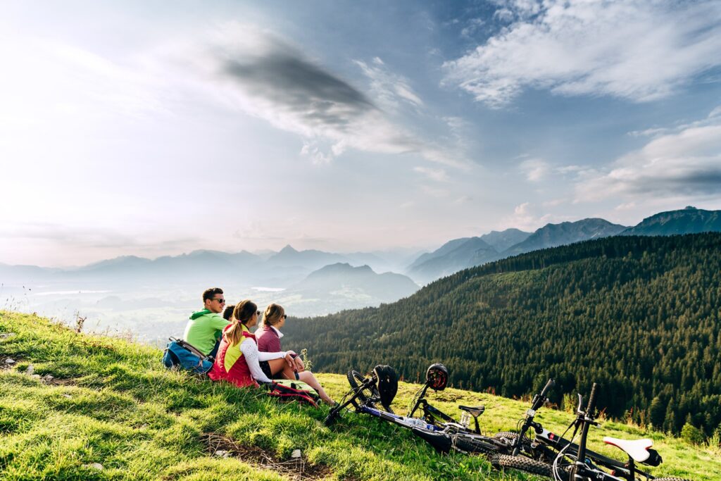 Four people in cycling gear are sitting on a green mountainside, looking down into the wooded valley. Their bikes are lying in the grass beside them.