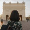 Eine Frau mit dunklen Haaren und einer Bluse mit Blumenmuster blickt mit dem Rücken zum Betrachter auf einen weißen, klassizistischen Triumphbogen, der im unscharfen Bokeh des Bildes noch gut zu erkennen ist.