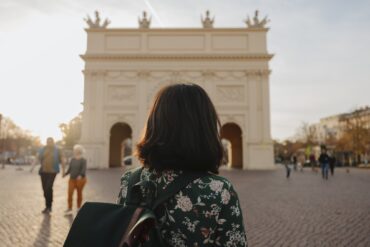 Eine Frau mit dunklen Haaren und einer Bluse mit Blumenmuster blickt mit dem Rücken zum Betrachter auf einen weißen, klassizistischen Triumphbogen, der im unscharfen Bokeh des Bildes noch gut zu erkennen ist.