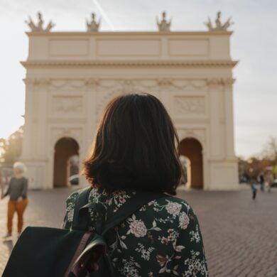 Eine Frau mit dunklen Haaren und einer Bluse mit Blumenmuster blickt mit dem Rücken zum Betrachter auf einen weißen, klassizistischen Triumphbogen, der im unscharfen Bokeh des Bildes noch gut zu erkennen ist.