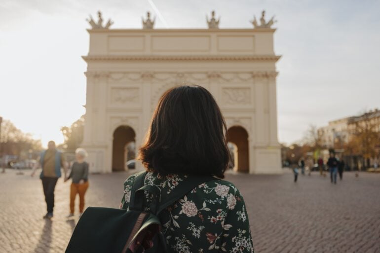 Eine Frau mit dunklen Haaren und einer Bluse mit Blumenmuster blickt mit dem Rücken zum Betrachter auf einen weißen, klassizistischen Triumphbogen, der im unscharfen Bokeh des Bildes noch gut zu erkennen ist.