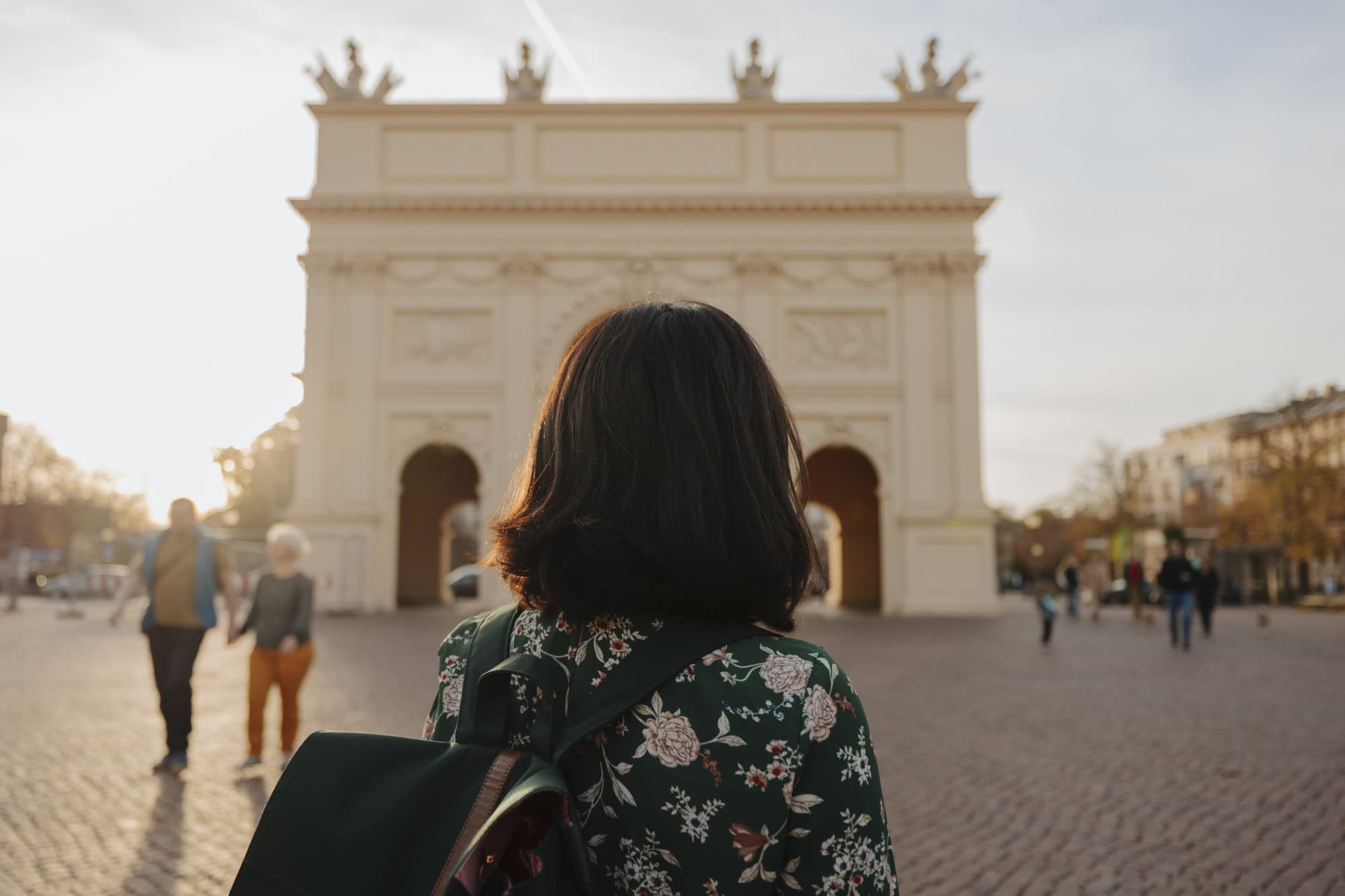 Eine Frau mit dunklen Haaren und einer Bluse mit Blumenmuster blickt mit dem Rücken zum Betrachter auf einen weißen, klassizistischen Triumphbogen, der im unscharfen Bokeh des Bildes noch gut zu erkennen ist.