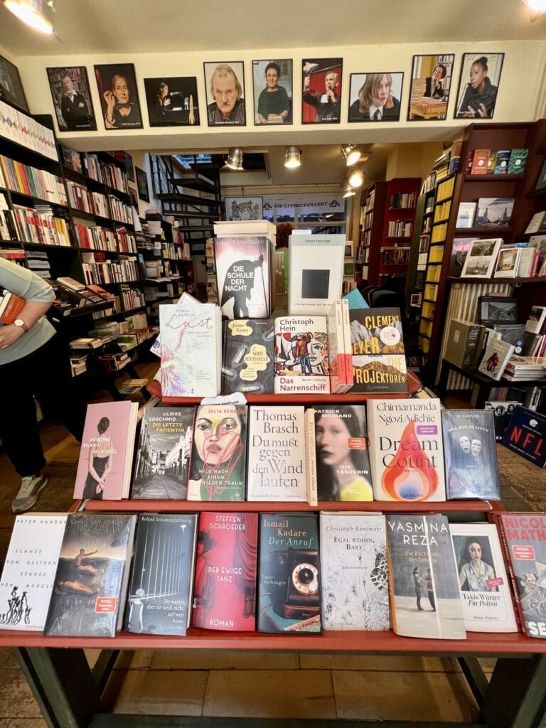 Ein in Form einer Pyramide angeordneter Produkttisch in einer Buchhandlung, mit aktuellen Bestsellern wie "Air" von Christian Kracht oder "Halbinsel" von Kristina Bilkau. Im Hintergrund mehrere Regale voll mit Büchern, links führt eine Wendeltreppe ins Obergeschoss.