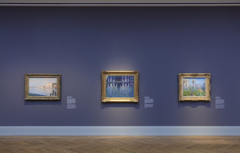 An einer anthrazitfarbenen Museumswand hängen nebeneinander drei Werke des Impressionismus in goldenen Rahmen.