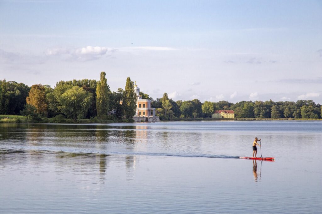 Ein Mann fährt auf einem roten SUP-Board über einen See. Das Ufer im Hintergrund ist mit vielen Bäumen bewachsen, mittig-links ragt ein zweigeschossiger Palas mit rundem Turmaufbau aus den Bäumen heraus.