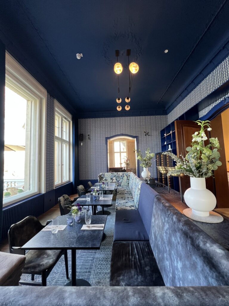 Blick in ein Restaurant; mittig drei Tische hintereinander, links jeweils ein Armlehnenstuhl und rechts eine lange Polstersitzbank mit blauen, gemusterten Polstern. Links im Bild zwei hohe Fenster, durch die viel Licht hineinfällt, rechts im Bild hinter der Bank eine Ablage und dahinter ein Regal. Im Hintergrund führt eine Tür in einen weiteren Raum. Auch die Wände des Raums sind in blau gehalten, mit vielen weißen Tupfern darauf.
