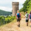 Drei Menschen in freizeitlicher Sommerkleidung wandern auf dem Kamm eines Weinbergs zur Linken in Richtung eines mittelalterlichen Rundturms. Unscharf im Hintergrund ein hoher Hügel mit Waldbewuchs.
