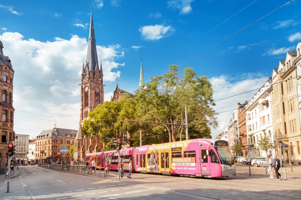 On a two-lane road, a tram with pink advertising prints is driving to the right out of the picture. Several people are standing at the stop on the opposite side. To the right behind the tram, a street with Wilhelminian-style apartment buildings leads into the background of the picture. In the center behind the tram are several deciduous trees, behind which a church tower rises into the sky. Everything seems very summery.