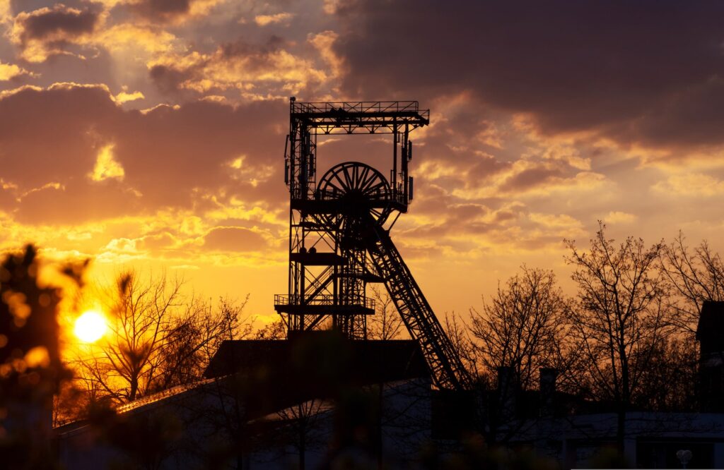 The silhouette of a mine tower with its characteristic conveyor wheel stands out against the bright setting sun. Around the tower are a few leafless trees, and the sky is colored gold.