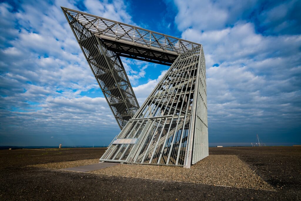 A work of art consisting of several wire struts, which initially extends diagonally upward to the right from the ground, then continues upward to the left at a 90-degree angle, and then curves sharply back down to the right behind the “start” in the foreground of the image. The sky behind it is blue with many clouds, and there is a flat concrete floor around the installation.
