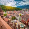 Eine mittelalterliche Stadt mit vielen roten Dächern und einem zentralen Marktplatz mit einer gotischen Kirche liegt in einem grünen Tal. Links im Bild prominent das Kirchendach. Hinter einem Berg im Hintergrund geht die Sonne unter.