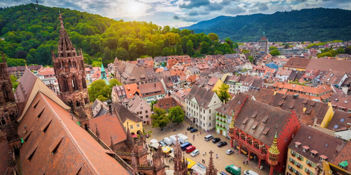 Eine mittelalterliche Stadt mit vielen roten Dächern und einem zentralen Marktplatz mit einer gotischen Kirche liegt in einem grünen Tal. Links im Bild prominent das Kirchendach. Hinter einem Berg im Hintergrund geht die Sonne unter.
