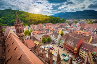 Eine mittelalterliche Stadt mit vielen roten Dächern und einem zentralen Marktplatz mit einer gotischen Kirche liegt in einem grünen Tal. Links im Bild prominent das Kirchendach. Hinter einem Berg im Hintergrund geht die Sonne unter.