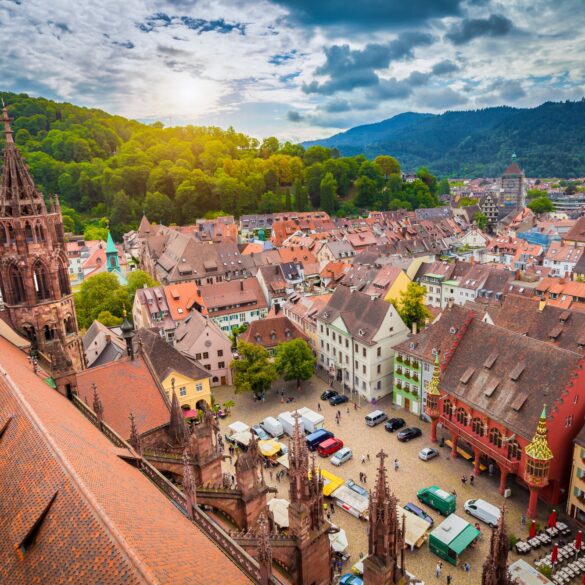 Eine mittelalterliche Stadt mit vielen roten Dächern und einem zentralen Marktplatz mit einer gotischen Kirche liegt in einem grünen Tal. Links im Bild prominent das Kirchendach. Hinter einem Berg im Hintergrund geht die Sonne unter.