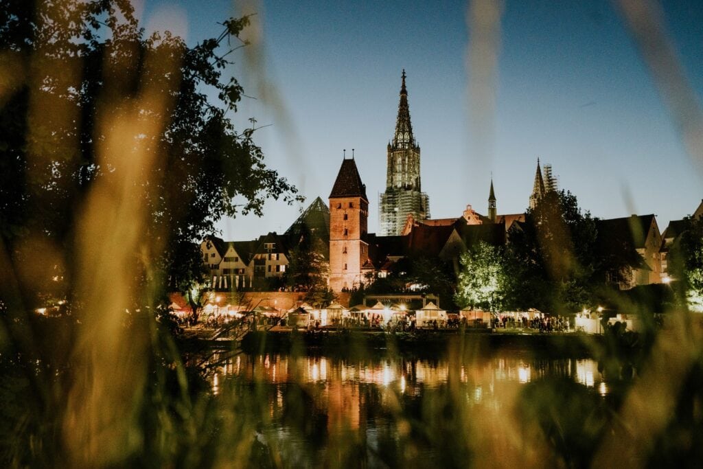 Blick auf eine mittelalterliche Stadt vom anderen Ufer eines Flusses am späten Abend. Mittig ein eckiger Turm, dahinter ein hoher, eingerüsteter Kirchenturm. Die gesamte Uferpromenade ist erleuchtet, einige Festzelte stehen darauf. Die Lichter spiegeln sich im still liegenden Fluss.