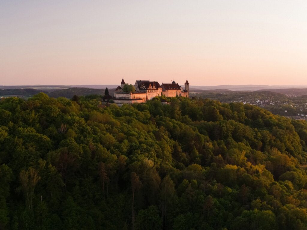Drone shot of a mighty castle rising from a dense forest on a hill above the landscape. The castle is bathed in red light from the setting sun, and the sky is clear.
