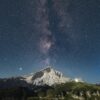 Über einem spitzen Berg inmitten einer Bergkette ist der Nachthimmel deutlich zu erkennen, die Milchstraße genau mittig über dem Gipfel.
