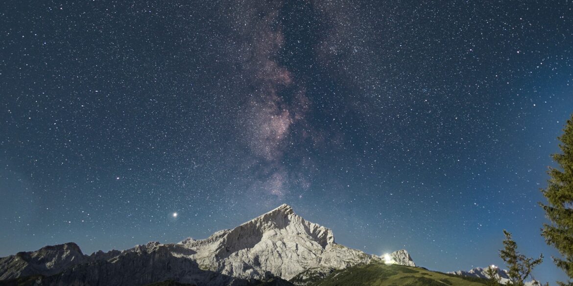 Über einem spitzen Berg inmitten einer Bergkette ist der Nachthimmel deutlich zu erkennen, die Milchstraße genau mittig über dem Gipfel.